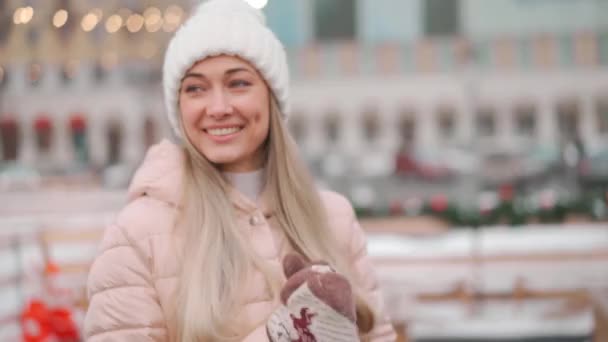 Vintertid. Jul semester, Ung vuxen kvinna njuter snöig vinter ute på stadens gata nära skridskoåkning rinken. Porträtt av kaukasisk vacker kvinna i vit stickad hatt på vintern — Stockvideo