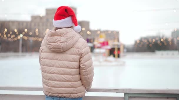 Kış zamanı. Noel tatili, Noel Baba şapkalı kadın buz pateni pistinin önünde duruyor sonra geriye bakıp gülümsüyor. Yılbaşı aydınlatma dekorasyonu. Kış tatili. — Stok video