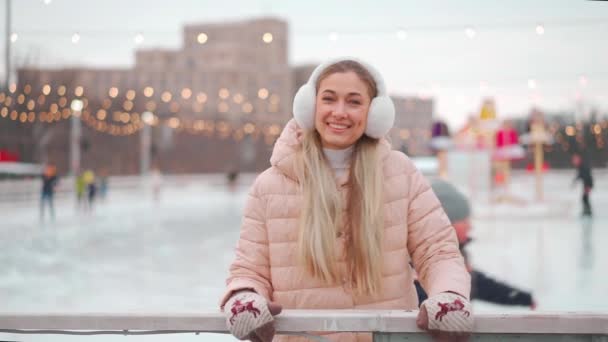 Invierno. Vacaciones de Navidad, mujer de pie fuera inclinarse en la cerca pista de hielo sonrisa e invitar a los amigos gesto de la mano. Fondo de decoración de iluminación navideña. Temporada de vacaciones de invierno — Vídeos de Stock