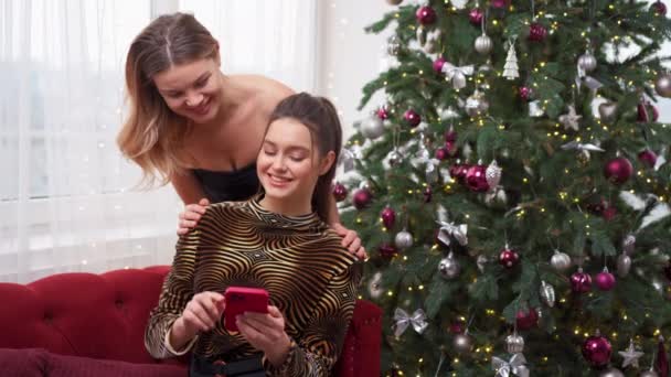 Deux femmes sont assises sur un canapé près de l'arbre de Noël regardant l'écran d'un smartphone discuter d'un profil sur un site de rencontres — Video