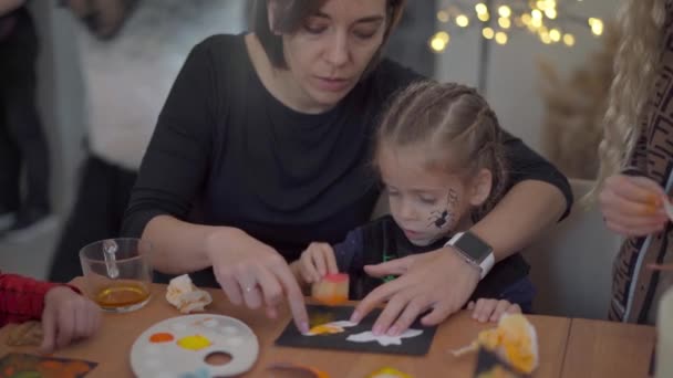 Маленька дівчинка одягнена в карнавальний костюм на Хелловін зі своєю матір'ю, сидить на столі малюнок ручної роботи на Хеллоуїн прикраси на вечірці — стокове відео