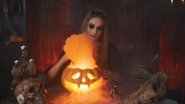 Halloween-konceptet. Svart häxa håller Halloween pumpa med snidade leende ansikte i handen sitter på bordet mörkt rum — Stockvideo