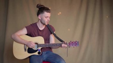 Akustik gitar çalan yakışıklı genç adam. Oturma odası kumaş arka planda.