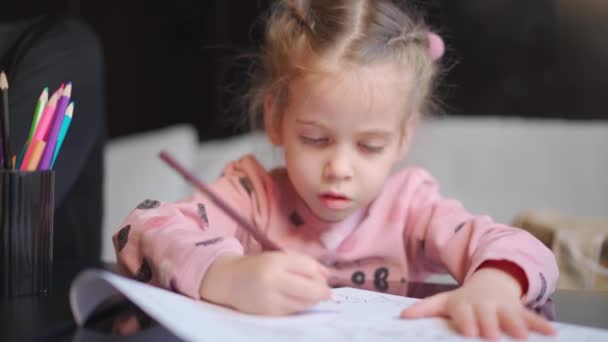 Lapsi piirustus istuu pöydällä keskellä ampua. Valkoihoinen pikkutyttö piirtää värillisillä kynillä. Nainen lapsi tekee kotitehtäviä — kuvapankkivideo