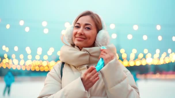 Förtroendeoptimistisk kvinna bära medicinsk skyddande ansiktsmask under covid 19 pandemi stående utanför vintern stad gata med julbelysning på bakgrunden ser kamera. — Stockvideo