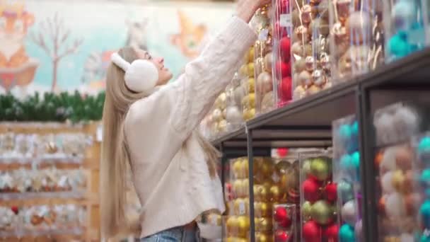 Shopping natalizio. La donna sceglie l'arredamento natalizio nel supermercato. Donna caucasica in cuffia di pelliccia vestita maglione bianco acquisto ornamento di Natale. Preparati per le vacanze invernali — Video Stock