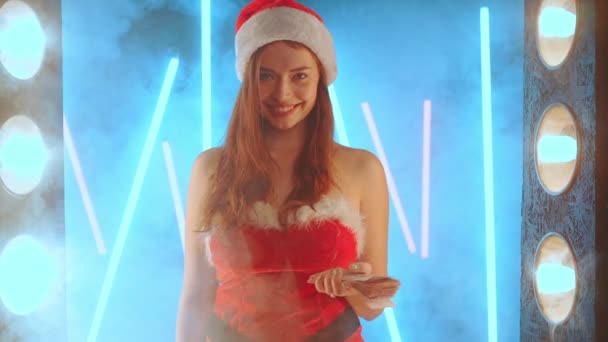 산타 모자를 쓰고 서서 네온 배경에 조명을 받으며 크리스마스 홀리데이 세일 컨셉의 여자가 돈을 던진다 — 비디오
