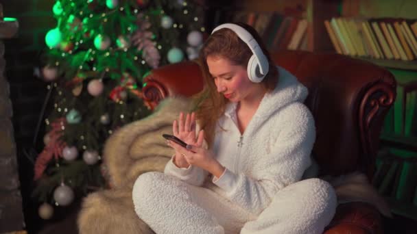 Νεαρή γυναίκα ακουστικά κάθεται δερμάτινη πολυθρόνα κοντά χριστουγεννιάτικο δέντρο κατέχει smartphone στο χέρι επιλέγει ένα τραγούδι — Αρχείο Βίντεο