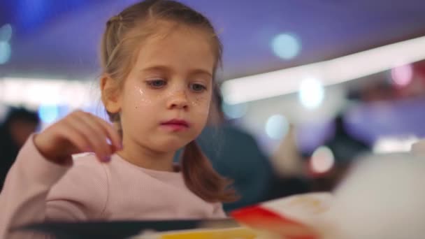 Niña con coletas comiendo papas fritas. Primer plano retrato caucásico femenino 5 años comer comida rápida en un restaurante por la noche con música ligera en el fondo — Vídeos de Stock