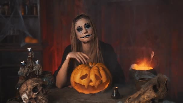 Halloween-konceptet. Svart häxa håller Halloween pumpa med snidade leende ansikte i handen sitter på bordet mörkt rum — Stockvideo
