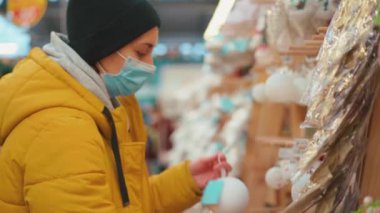 Sarı kışlık ceketli bir kadın alışveriş merkezinde Noel süslerini seçiyor. Tıbbi koruyucu maskeli yetişkin beyaz kadın yeni yıl süsü alıyor. 