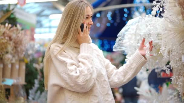Kerstinkopen, Europese vrouw kiest takken voor kerstkrans indoor in supermarkt praten aan de telefoon. Blanke vrouw kopen kerstdecor op vakantie markt — Stockvideo
