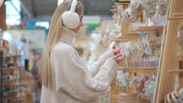 Kerstinkopen. Positieve Kaukasische vrouw wandelen supermarkt kiezen voor kerst ornament, gekleed witte trui en bont koptelefoon. Volwassen vrouw 30 jaar kerstdecor kopen — Stockvideo