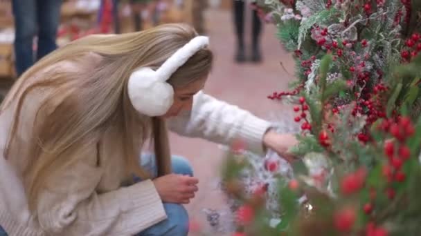 Kerstinkopen, Europese vrouw kiest takken voor kerstkrans indoor in supermarkt. Blanke vrouw kopen kerstdecor op vakantie markt, gekleed bont koptelefoon en witte trui. — Stockvideo