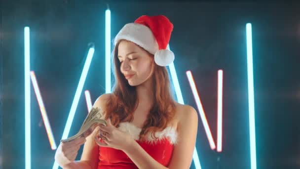 Kerstmis Vakantie verkoop concept Vrouw gekleed in Santa kostuum S Santa hoed met geld in de hand, terwijl staande verlicht neon achtergrond — Stockvideo