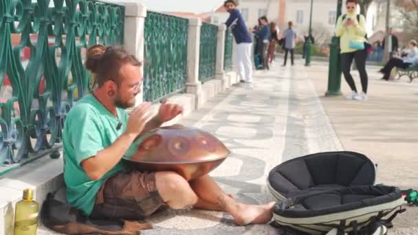 Portugal, Lisboa Março 2022 músico de rua de cabelos compridos tocando tambor pendurado na praça do mercado. Sentado descalço no cobertor. pessoas andando em segundo plano. — Vídeo de Stock