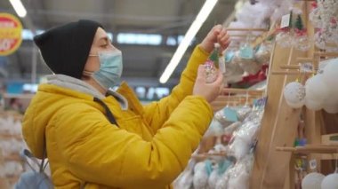Sarı kışlık ceketli bir kadın alışveriş merkezinde Noel süslerini seçiyor. Tıbbi koruyucu maskeli yetişkin beyaz kadın yeni yıl süsü alıyor. 