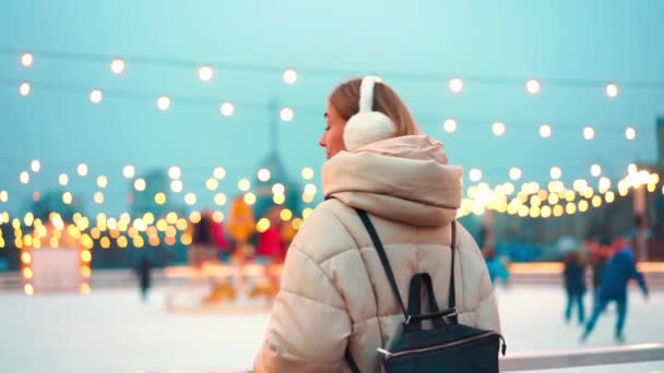 Wintertime. Christmas holiday evening, woman standing outside looking ice rink than look back and smile. Christmas illumination and decoration background. Winter holiday season — Stock Video
