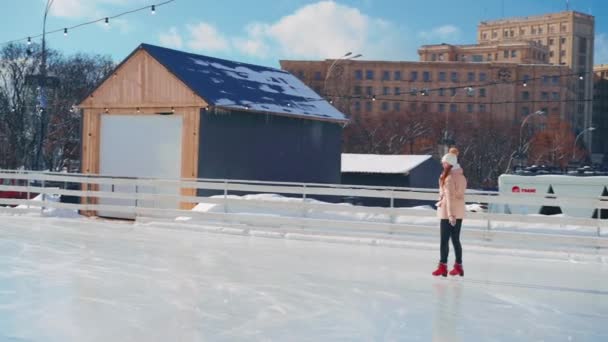 Joven mujer sonriente patinaje sobre hielo fuera en pista de hielo plaza central de la ciudad en las vacaciones de Navidad, ocio activo de invierno en un día cálido y soleado. Efecto de mano. Amateur caucásico hembra patinador de hielo — Vídeos de Stock