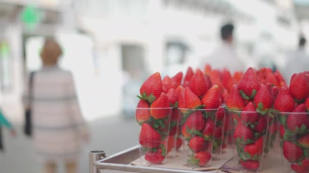 Свежие клубничные фрукты в пластиковых чашках на рынке. Открытый фруктово-ягодный рынок. Размытые люди на фоне городской улицы. — стоковое видео