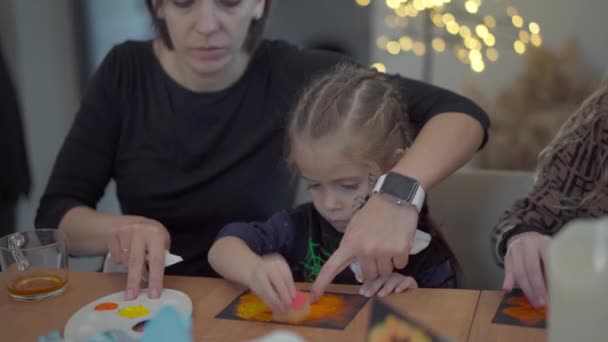 Klein meisje gekleed Halloween carnaval kostuum met haar moeder zitten op de tafel tekening handgemaakte Halloween decoratie op feest — Stockvideo