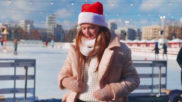 Νεαρή χαμογελαστή γυναίκα Σάντα καπέλο παγοδρομία έξω στο παγοδρόμιο ντυμένο λευκό πουλόβερ. Χριστουγεννιάτικες διακοπές, δραστήριος χειμερινός ελεύθερος χρόνος. Ερασιτέχνης καυκάσιος γυναικάς παγοδρόμος — Αρχείο Βίντεο