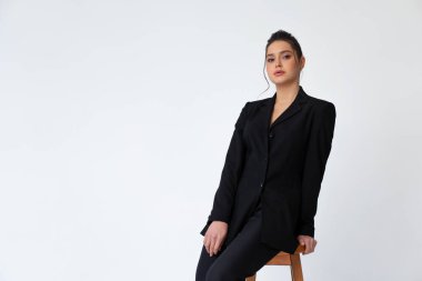 Genç, modaya uygun bir iş kadını yüksek bar ahşap koltukta oturuyor. Siyah ofis elbisesi giyip beyaz arka planda izole edilmiş. Beyaz kadın iş adamı. Kameraya bakarken dalgın.