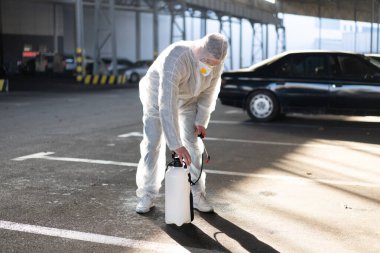 Beyaz elbiseli bir adam karantina sırasında yüzey antibakteriyel dezenfektan püskürtücü sıkarak ekipmanları hazırlıyor pompayı pompaya pompalıyor.