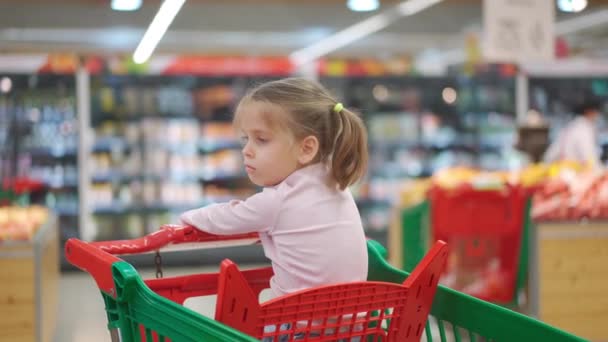 Bambino, bambina 5 anni seduta carrello della spesa nel supermercato mentre la madre sceglie le merci per comprarlo Femmina bambino carrello della spesa negozio di alimentari o supermercato. Bambino che va a fare shopping. — Video Stock