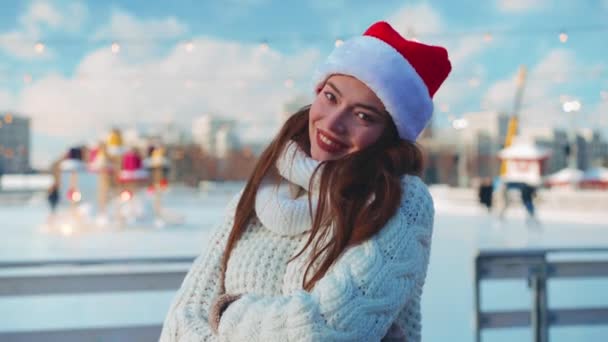 Νεαρή χαμογελαστή γυναίκα Σάντα καπέλο παγοδρομία έξω στο παγοδρόμιο ντυμένο λευκό πουλόβερ. Χριστουγεννιάτικες διακοπές, δραστήριος χειμερινός ελεύθερος χρόνος. Ερασιτέχνης καυκάσιος γυναικάς παγοδρόμος — Αρχείο Βίντεο