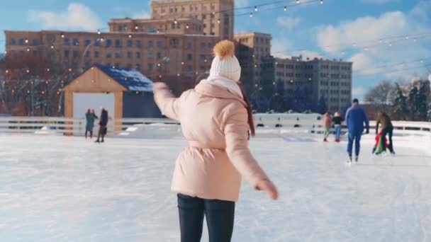 Joven mujer sonriente patinaje sobre hielo fuera en pista de hielo plaza central de la ciudad en las vacaciones de Navidad, ocio activo de invierno en un día cálido y soleado. Efecto de mano. Amateur caucásico hembra patinador de hielo — Vídeos de Stock