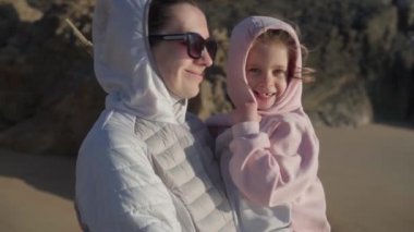 Anne, baharda çocuğu okyanus kıyısında elinde tutuyor, sıcak kıyafetler giyiyor ve video efekti uyguluyor.