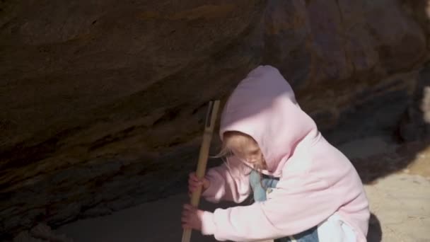 Niño jugando en la playa cerca de la roca, niña está cavando arena con palo como un arqueólogo caucásico mujer 5 años de edad, vestida con capucha rosa divertirse jugando en la playa — Vídeos de Stock
