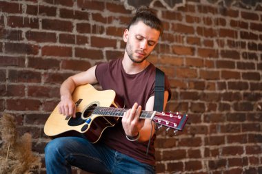 Akustik gitarlı, tuğla duvarlı, müzik çalan, hayatın tadını çıkaran yakışıklı beyaz gitarist ev yapımı, müzikal enstrüman çalıyor.