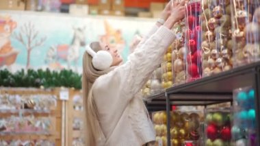 Noel alışverişi. Kadınlar süpermarkette Noel dekorunu seçerler. Kürk kulaklıklı beyaz kazaklı beyaz bir kadın Noel süsü alıyor. Kış tatiline hazırlanın.