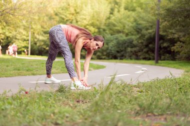 Atletik kadın, yaz parkında koşu antrenmanlarından önce bacaklarını ısıtıyor. Sabah kardiyo antrenmanı. Orta yaşlı, beyaz bir kadın cesedi ısıtıyor.