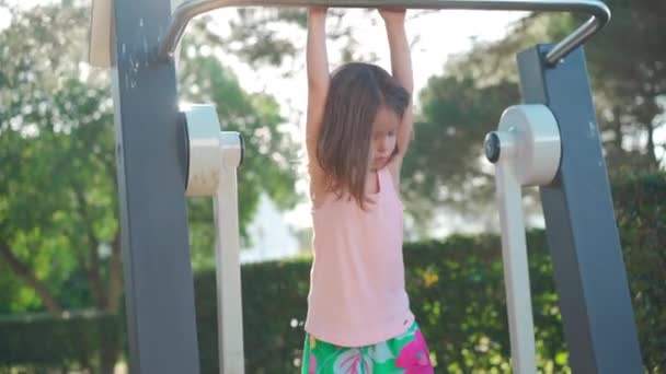 Active little girl, child, trains on simulator city park. She pulls and pushes body. Muscle strength improvement during holidays. Caucasian little girl 5 years active sport leisure outdoors — ストック動画