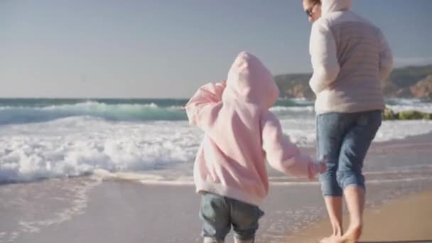 어머니를 위해 달려가는 아이는 봄에 따뜻 한 옷을 입고 따뜻 한 맨발의 손으로 비디오 효과를 내면서 바다 해변에서 노는 모습을 따라잡으려고 한다 — 비디오
