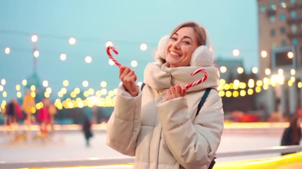 Mujer en orejeras de pie al aire libre cerca de pista de patinaje sobre hielo en la plaza central de la ciudad en la noche de vacaciones de invierno. Gente patinando sobre fondo con luz navideña. — Vídeos de Stock