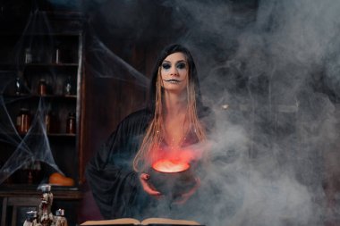 Cadılar Bayramı, cadı sihirli kitap ve kazan kullanın zehir ya da aşk iksiri hazırlayın. Kara büyü okült kadın büyücü duman ve örümcek ağıyla karanlık odada. Büyücü sihirli iksir pişiriyor. Kapat.