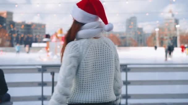 Молода усміхнена жінка Санта-капелюх катається на ковзанах, одягнених у білий светр. Різдвяні свята, активне зимове дозвілля Ручний ефект. аматорська біла жінка з льодом — стокове відео