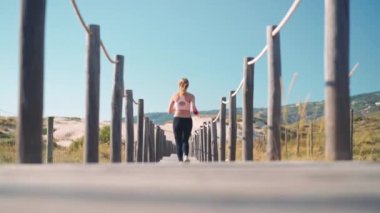 Plaj ahşap patikasında koşan atletik sporcu kadın yavaş çekim takip ediyor. Sevimli, genç, beyaz kadın deniz kenarında koşuyor. Sağlıklı yaşam tarzının geriden görünüşünün tadını çıkarıyor.. 