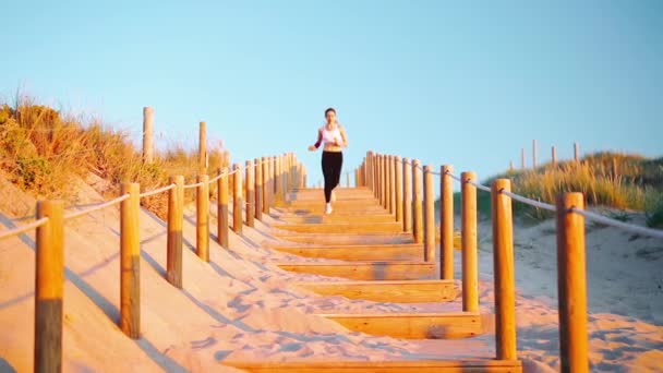 ビーチ木製の階段のパススローモーションで実行されているアスレチックスポーツ女性。海辺で健康的なライフスタイルを楽しむ若い大人の白人女性のジョギング屋外 — ストック動画