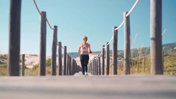 Desportista atlética correndo na praia caminho de madeira câmera lenta rastreamento tiro. Adorável jovem adulto caucasiano feminino jogging ao ar livre à beira-mar desfrutando de estilo de vida saudável vista traseira. — Vídeo de Stock