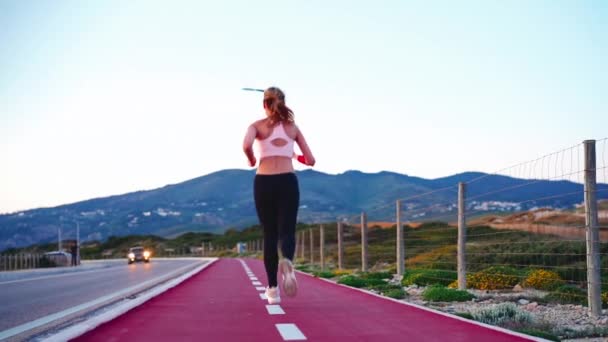 Dağın arka planında, yolun kenarında bisiklet yolunda koşan uzun boylu atletik bayan koşucu. Sportif kadın doğa koşusu. Sağlıklı yaşam tarzı — Stok video