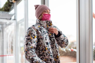 Pembe maskeli Avrupalı kadın kışlık giysileri karantinaya, koronavirüs salgınına, alışverişe, dükkana, sokağa taşıdı. Şehir caddesindeki pencereden bakan kışlık ceketli.