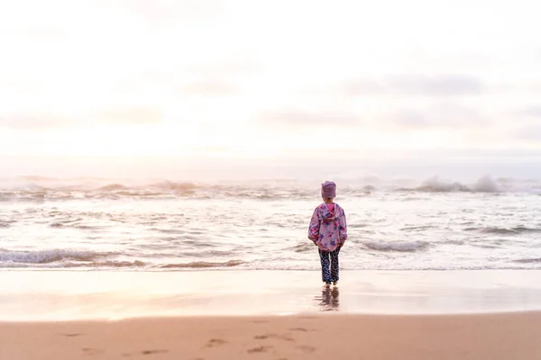 孤独な少女は 海のビーチに戻るカメラには 日の出を見て立っている 孤独小児うつ病 白人女性の子供5年は暖かい服を着て日没の海のビーチを歩く — ストック写真