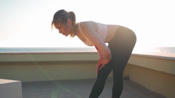 女跑步者靠着膝盖在美丽的日落背景下休息 白种人的女慢跑者在晚上锻炼后 穿着运动服站着休息 健康生活方式概念 — 图库视频影像