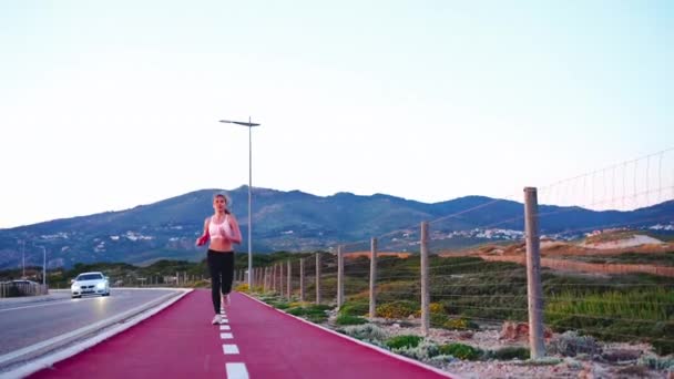 山の背景を持つ道路の近くに自転車レーンを実行しているフル長さのビューのアスレチック女性ジョガー スポーティな女性のジョギングの性質 健康的なライフスタイルのコンセプト スポーツウェア 夕方のトレーニング — ストック動画