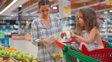 Kızı süpermarket arabasında oturuyor. Sevimli aile, anne ve küçük kız hipermarketten meyve ve sebze alıyorlar. Vejetaryenlik ve sağlıklı beslenme konsepti Aile bakkal alışverişi yeşil elma